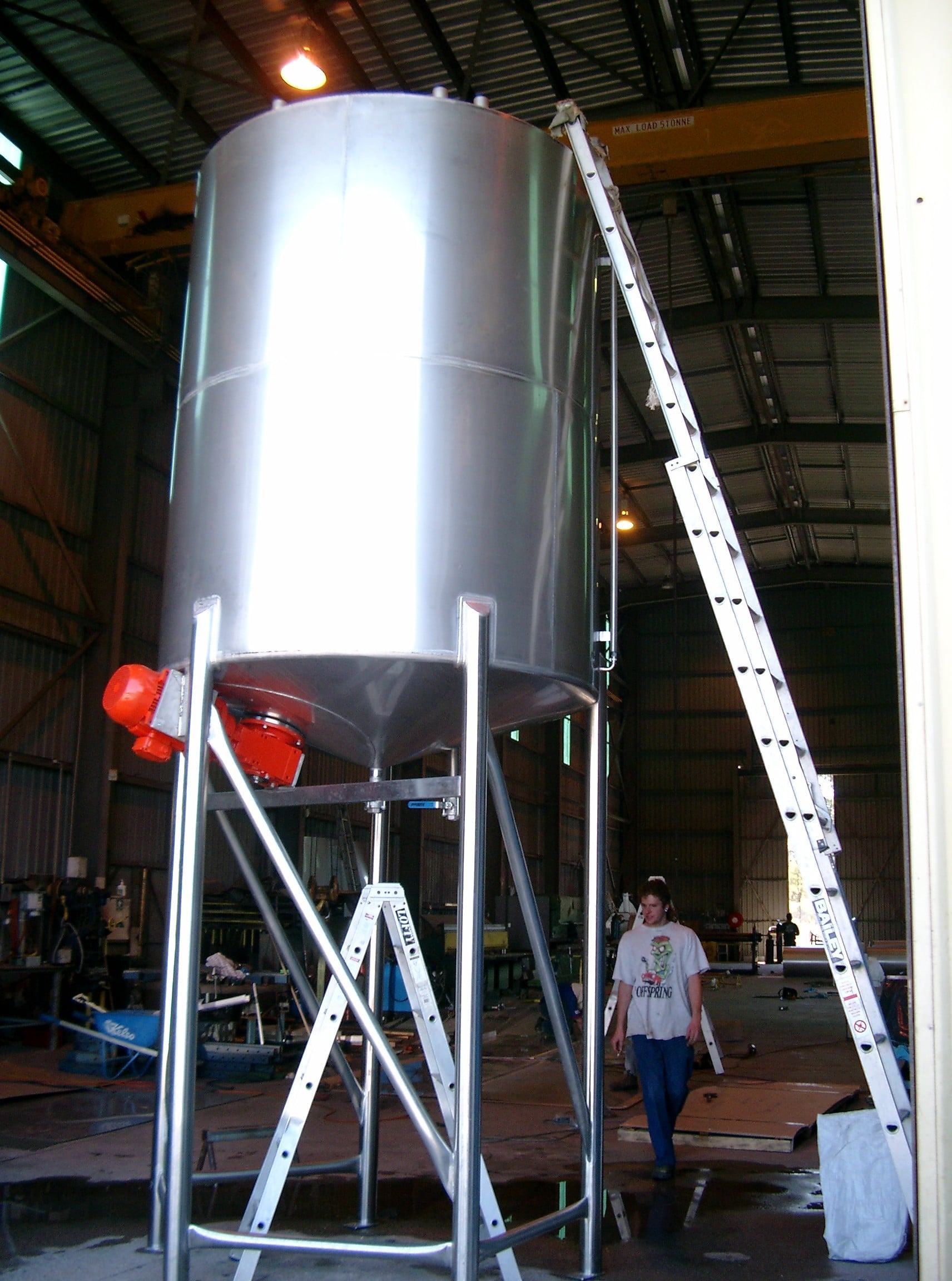 A stainless steel tank with conical base, with a base entry mixer mounted to it. A ladder leans against the tank for scale. Stainless legs and frame are visible